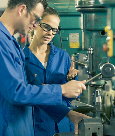 Vergrößerungsansichten für Bild: Ein Mann und eine Frau stehen in Arbeitskleidung an einer Maschine. Sie tragen Schutzbrillen.
