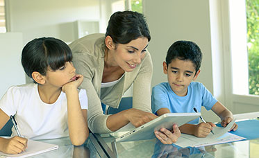 Das Bild zeigt zwei Kinder, die an einem Tisch sitzen und von einer jungen Frau Hilfe bei den Hausaufgaben bekommen.