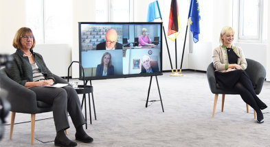Frau Wenng, Moderatorin (links) und Frau Staatsministerin Carolina Trautner (rechts) in Stühlen sitzend. Dazwischen ein Bildschirm mit vier eingeblendeten weiteren Personen, die digital zugeschaltet sind.