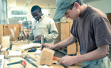 Vergrößerungsansichten für Bild: Zwei junge Männer arbeiten gemeinsam in einer Tischlerei. Sie tragen beide Arbeitskleidung und stehen an einer Werkbank, auf der Arbeitsmaterialien liegen.