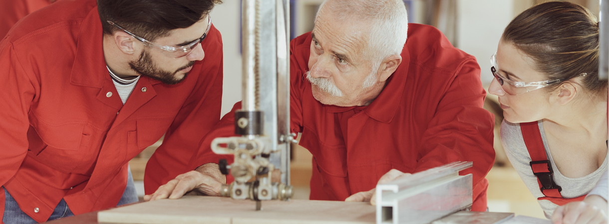 Drei Personen unterschiedlichen Alters arbeiten in Arbeitskleidung an einer Holzschneidemaschine. 