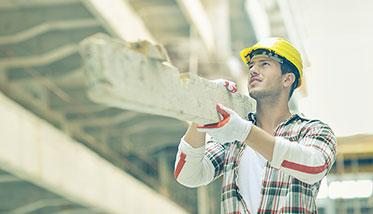 Ein junger Mann mit Helm und Schutzhandschuhen trägt auf seiner rechten Schulter einen Betonstein. Im Hintergrund befindet sich eine Baustelle.
