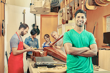 Ein lachender junger Mann mit Migrationshintergrund schaut in die Kamera, hinter ihm arbeiten ein paar Menschen an einem Instrument