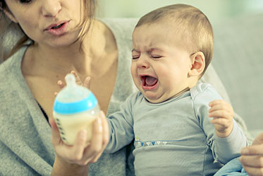 Frau mit schreiendem Baby auf dem Schoß und Babyflasche in der Hand