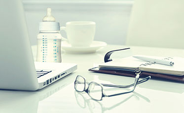 Nahaufnahme: Auf einem Tisch steht ein Laptop neben einer Babyflasche und einer Tasse. Außerdem liegen eine Brille und ein aufgeschlagenes Notizbuch samt Kugelschreiber auf dem Tisch