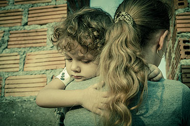 Nahaufnahme: Eine Frau ist von hinten zu sehen mit einem Kind auf dem Arm, das traurig über ihre Schulter schaut