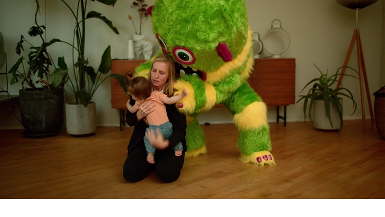 Eine Frau beschützt ein Kleinkind vor einem übergroßen grün-gelb gestreiften Plüschmonster.