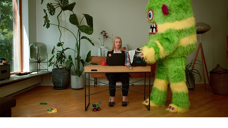 Ein übergroßes grün-gelb gestreiftes Plüschmonster stellt einen vollen Wäschekorb auf einen Schreibtisch, an dem eine Frau sitzt.