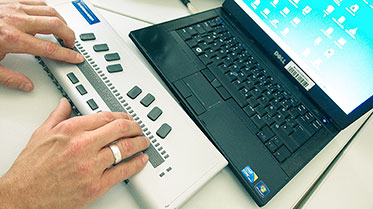 Vergrößerungsansichten für Bild: Notebook mit angeschlossenem Braille-Display. Die Hände des Benutzers liegen auf der Braille-Zeile.