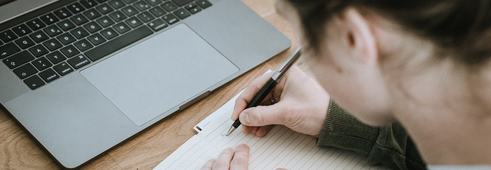 Person schreibt in einem notizbuch etwas auf. Vor der Person steht ein Notebook in silber mit schwarzer Tastatur. 