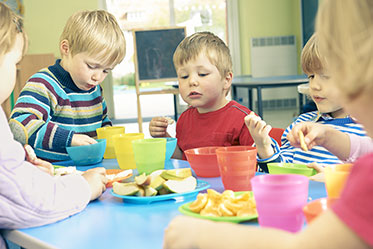 Vergrößerungsansichten für Bild: Nahaufnahme: Mehrere Kinder sitzen an einem Tisch und essen Obst in Form von klein geschnittenen Äpfeln und Mandarinen.