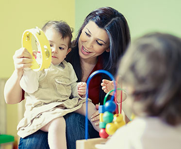 Nahaufnahme: Mitarbeiterin einer Kinderkrippe beim Spielen mit einem kleinen Kind, das mit einer Rassel auf ihrem Schoß sitzt. 