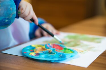 Kind mit Malschürze malt mit einem Pinsel, vor ihm ein Blattpapier und eine Farbmischpalette auf einem Tisch liegend 
