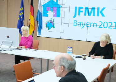 Staatsministerin Carolina Trautner mit pinkem Oberteil vor einem großen Bildschirm. Im Hintergrund eine Bildschirmleinwand mit eingeblendetem Logo der JFMK Bayern 2021