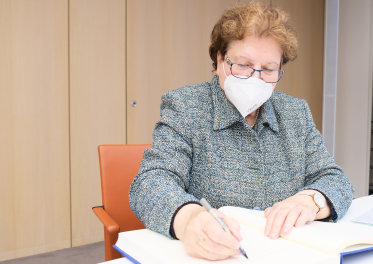 Landtagspräsidentin a.D. Barbara Stamm mit grau gesprenkelten Oberteil und Maske unterschreibend sitzend an einem Tisch