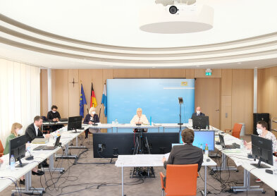Auf dem Bild ist ein großer Raum mit Blick auf eine hellblaue Pressewand zu sehen. Darauf unter anderem Staatsministerin Carolina Trautner sitzend an einem Tisch vor zwei großen Bildschirmen.