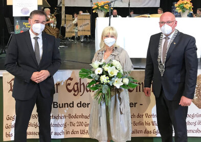 Auf diesem Bild sind drei Personen mit Maske zu sehen. In der Mitte Staatsministerin Carolina Trautner mit einem großen Blumenstrauß in den Armen. 