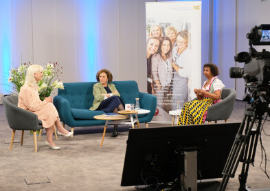 Auf diesem Bild sind drei Frauen sitzend auf zwei grauen Sesseln mit Holzfüssen und einem petrolfarbenem Sofa. In der Mitte steht ein holzfarbener-weißer Tisch und der Hintergrund ist blau beleuchtet. Im rechten Bildrand ist Technikequipment vom Studio zu sehen.