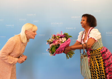 Auf diesem Bild ist Staatsministerin Carolina Trautner sowie der Gast des Abends zu sehen. Beide stehen vor einer hellblauen Wand. Der Gast trägt ein grün-rosanes Dirndl mit weißer Bluse. Sie übergibt einen Blumenstrauß. 