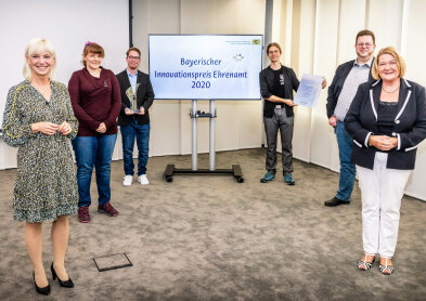 Auf diesem Bild sind sechs personen zu sehen. Sie lächeln in die Kamera. Hinten in der Mitte ist ein Bildschirm auf dem Steht Bayerischer Innovationspreis Ehrenamt 2020