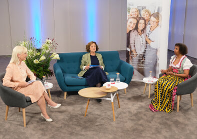 Auf diesem Bild sind drei Frauen sitzend auf zwei grauen Sesseln mit Holzfüssen und einem petrolfarbenem Sofa. In der Mitte steht ein holzfarbener-weißer Tisch und der Hintergrund ist blau beleuchtet. 