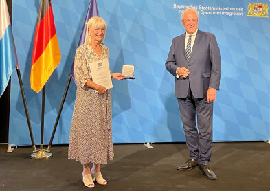 Links steht Sozialministerin Carolina Trautner mit einer Urkunde und einer Medialle in der HAnd. REchts Innenminister Joachim Herrmann. Dahinter eine blaue Pressewand und drei Flaggen von Europa, Deutschland und Bayern. 