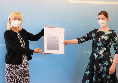 Staatsministerin Carolina Tratuner und Dr. Annette Greiner vor hellblauem Hintergrund während der Überreichung einer Urkunde.