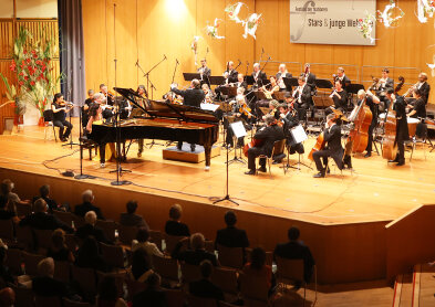 Auf diesem Bild ist eine Bühne von oben zu sehen. Darauf sitzen ca. 20 Personen eines Orchesters mit ihren Instrumenten. Im Vorderdrung ein Flügel. 
