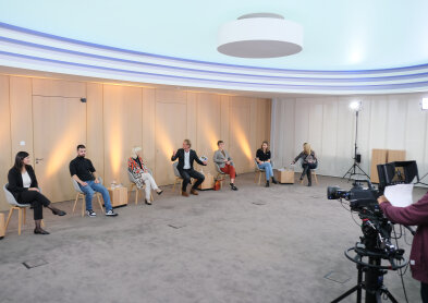 Auf dem Bild sind 7 Personen zu sehen. Sie sitzen in einer Reihe vor einem hellen hölzernen Hintergrund auf grauen modernen Stühlen. Rechts im Bild ist eine Kamera angeschnitten. 