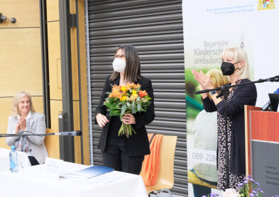 Sozialministerin Carolina Trautner klatschend neben einer Frau mit Blumen in der Hand. 