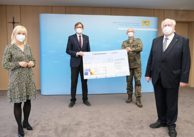 Staatsminiterin Carolina Trautner mit drei weiteren Herren auf dem Bild. Aufgestellt vor einer hellblauen Wand. Die beiden HErren in der Mitte halten einen Überdimensionierten Check in der Hand. 