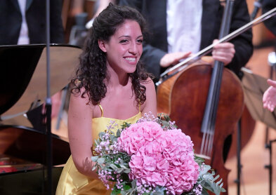 Auf dem Bild ist eine Dame im gelben Kleid zu sehen. Sie hält einen Blumenstrauß mit großen pinken Blüten und lächelt. 