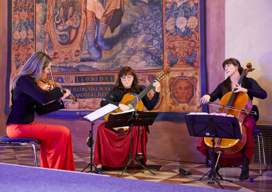 Auf dem Bild sind 3 Damen mit Musikinstrumenten zu sehen. Links eine Geige in der Hand, in der Mitte eine Gitarre und rechts ein Cello.. 