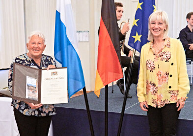 Auf dem Bild sind zwei Personen zu sehen. links eine Arbeitsjubilarin mit Urkunde in der HAnd und rechts Arbeitsministerin Carolina Trautner. Sie stehen vor drei Fahnen, der Bayern-, der Deutschland- und der Europafahne. 