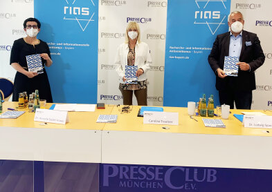 Auf dem Bild ist Frau Staatsministerin Carolina Trautner zusammen mit zwei weiteren Personen vor weißen und blauen Pressewänden zu sehen.