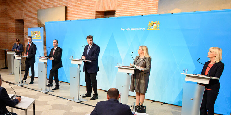 Ministerpräsident Markus Söder und vier weitere Mitglieder der Bayerischen Staatsregierung stehen vor einer blauen Pressewand. Alle Personen stehen hinter einem Rednerpult mit Mikrofon.
