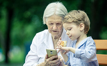 Nahaufnahme: Eine ältere Frau sitzt mit ihrem Enkel auf einer Bank und beide schauen sich etwas auf einem Smartphone an