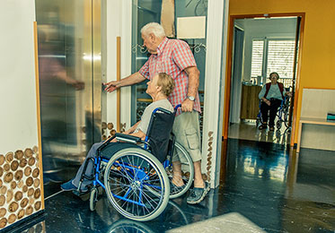 Älterer Mann und Frau im Rollstuhl vor einem Aufzug.