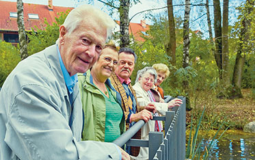 Mehrere ältere Menschen auf einer Terrasse an einem Weiher; im Hintergrund Parklandschaft.