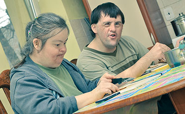 Eine Frau und ein Mann mit Behinderung sitzen an einem Tisch. Sie haben jeweils einen Stift in der Hand und einen Papierblock vor sich. 