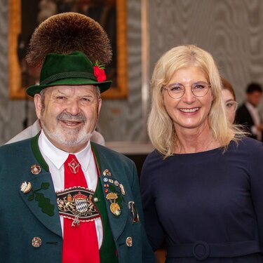 Das Foto zeigt Bayerns Sozialministerin Ulrike Scharf neben einem Ehrenamtlichen in feierlicher Tracht