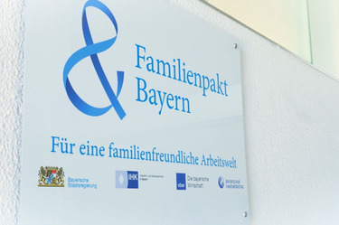 Auf dem Bild ist ein wei&szlig;es Schild bzw. Emblem zu sehen. Darauf ist ist blauer Schrift &quot;Familienpakt Bayern F&uuml;r eine familienfreundliche Arbeitswelt&quot; zu lesen.